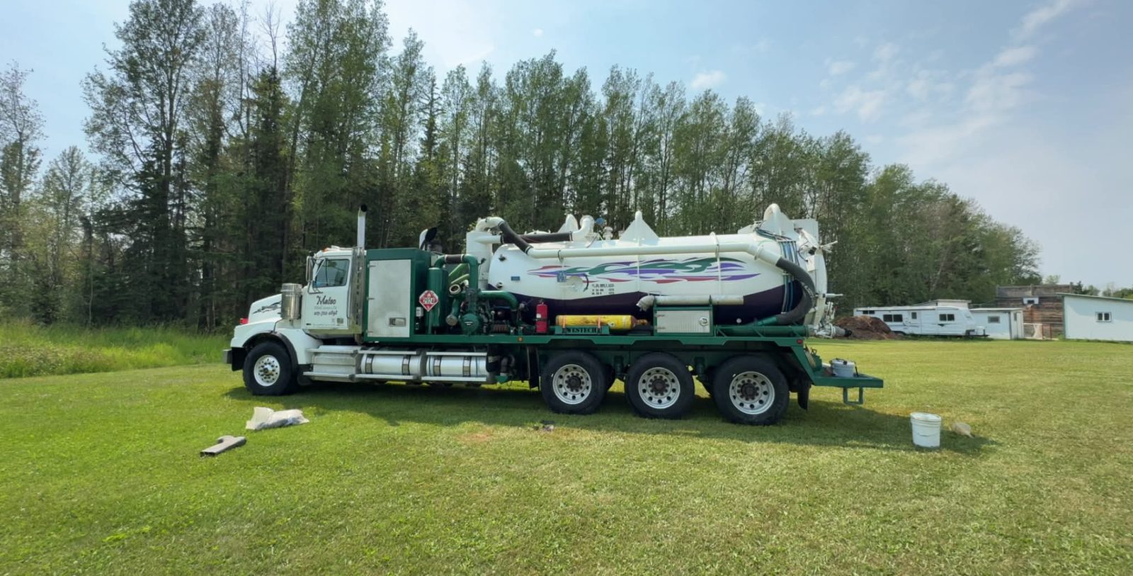 Side view of a white Mateo Western Star vacuum truck with a green deck parked on green grass in front of spruce trees